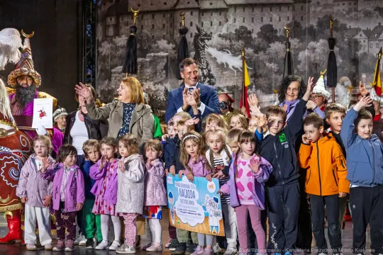 Grupa dzieci i dorosłych na scenie, wszyscy uśmiechnięci i klaskający.