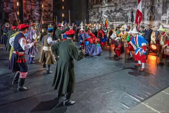Scena z historycznego przedstawienia z postaciami w strojach z epoki, trzymającymi broń i flagi.