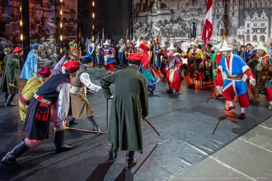 Scena z przedstawienia teatralnego z postaciami w historycznych strojach, tańczących i trzymających broń.