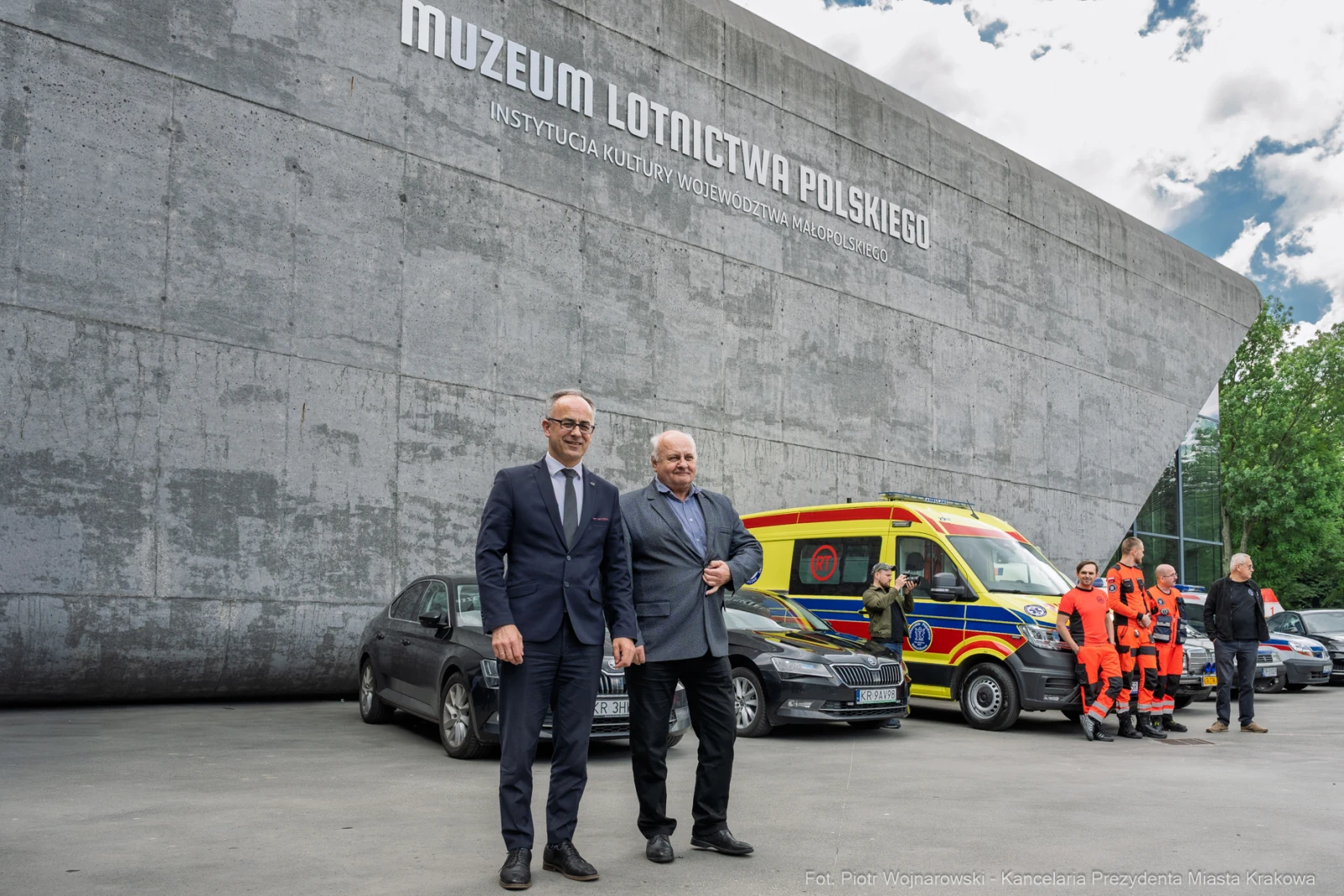 Dwaj mężczyźni w garniturach przed nowoczesnym budynkiem muzeum, w tle karetka i pojazdy ratunkowe.  Autor: Piotr Wojnarowski