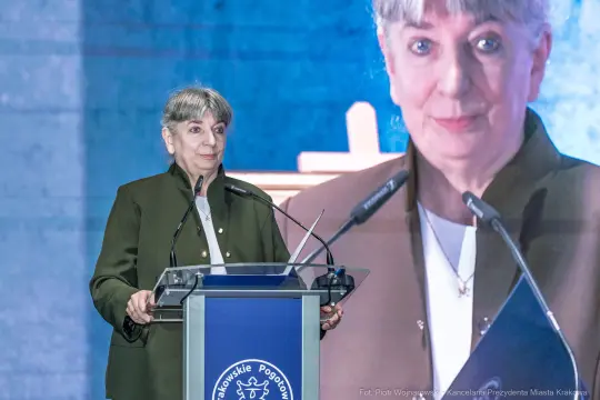 Kobieta przemawia z mównicy, w tle widoczny ekran z jej wizerunkiem.