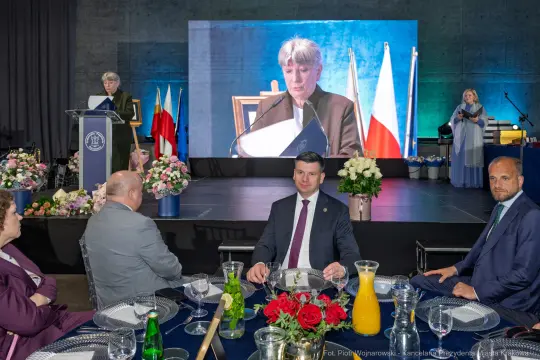 Uroczystość z przemówieniem, goście przy stole, ekran z prezentacją.