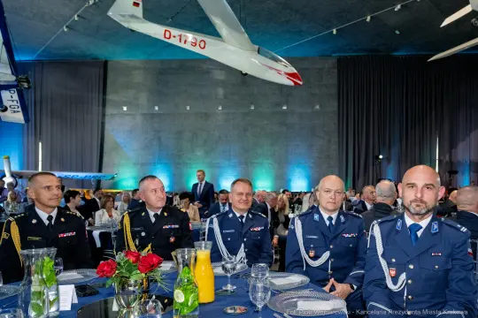 Grupa mężczyzn w mundurach siedzących przy eleganckim stole w sali z samolotem w tle.