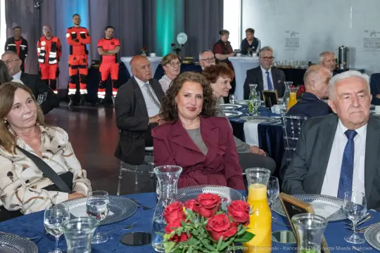 Grupa osób siedzących przy eleganckim stole, w tle strażacy w mundurach.