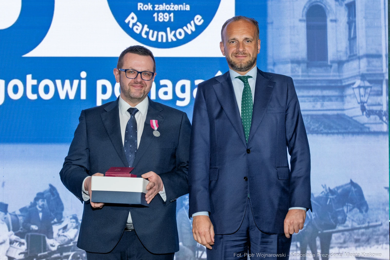 Dwaj mężczyźni na uroczystości, jeden z medalem, drugi z prezentem. Tło z logo i datą.  Autor: Piotr Wojnarowski