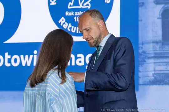 Mężczyzna w garniturze rozmawia z kobietą w niebieskiej bluzce w kontekście wydarzenia publicznego.