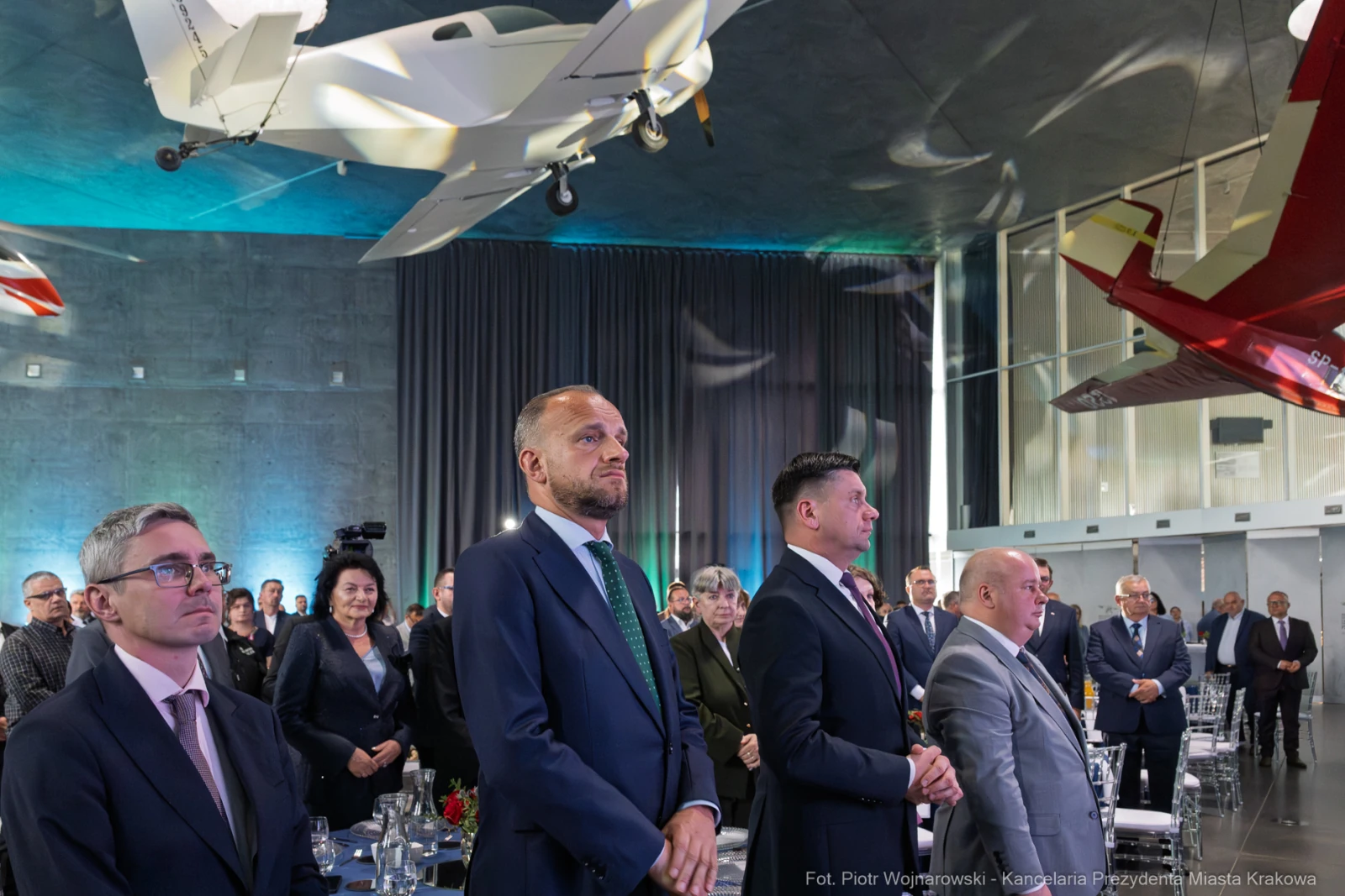 Grupa ludzi w eleganckich strojach podczas wydarzenia w nowoczesnej sali z modelami samolotów.  Autor: Piotr Wojnarowski