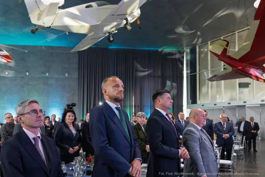 Grupa ludzi w eleganckich strojach podczas wydarzenia w nowoczesnej sali z modelami samolotów.