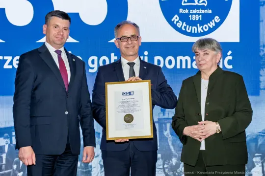 Trzech mężczyzn i kobiet w formalnych strojach trzymających dyplom na tle niebieskiego tła.