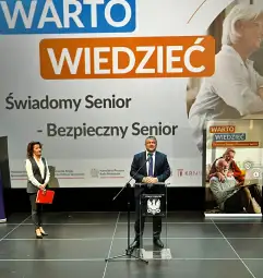 Konferencja o bezpieczeństwie seniorów z prelegentami i prezentacją na tle.