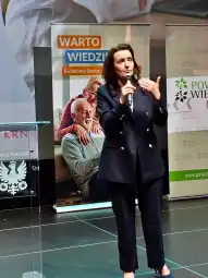 Kobieta w eleganckim stroju przemawia na tle bannerów promujących świadomość seniorów.