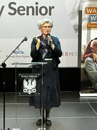 Kobieta w okularach i z siwymi włosami przemawia do publiczności.