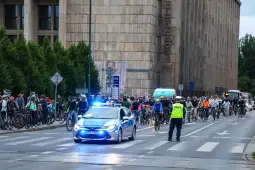 Grupa rowerzystów zatrzymana przez policję na ulicy, w tle radiowóz z włączonymi sygnałami świetlnymi.