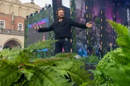 Mężczyzna z mikrofonem na scenie, otoczony zielenią, w tle widoczne dekoracje i publiczność.