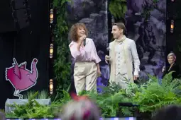 Dwoje artystów występuje na scenie, otoczeni zielenią i dekoracjami.