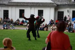 Dwaj performerzy w kostiumach tańczą na trawie przed publicznością.