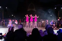 Trzy tancerki w różowych sukienkach występują na scenie przed publicznością podczas deszczu.
