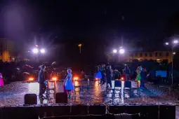 Występ artystyczny na deszczowej scenie z oświetleniem i widownią z parasolami.
