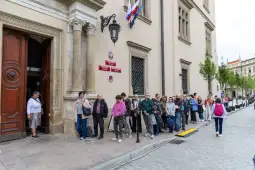 Ludzie stoją w kolejce przed budynkiem z flagami i tablicami informacyjnymi.