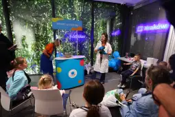 Kobieta w białym fartuchu prowadzi zajęcia dla dzieci w kolorowym otoczeniu.