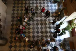 Grupa ludzi stojąca w okręgu na szachownicowej podłodze, otoczona roślinami.