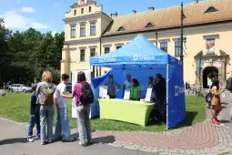 Ludzie przy stoisku informacyjnym w Krakowie, zielony namiot, otoczenie parku.