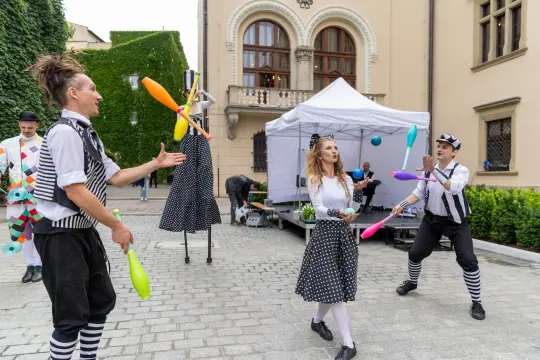 Cyrkowcy żonglują kolorowymi przedmiotami na tle budynku i namiotu.