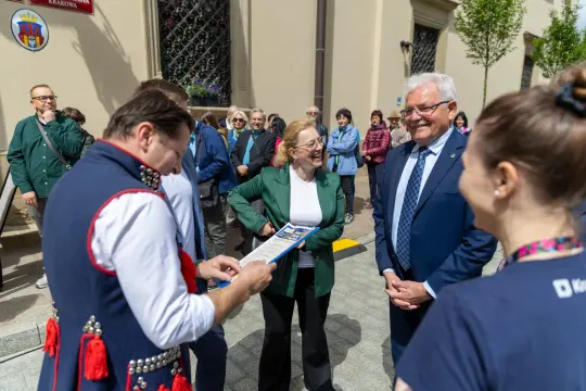 Grupa ludzi rozmawia na ulicy, w tle budynek i drzewa.