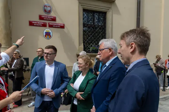 Cztery osoby w formalnych strojach rozmawiają przed budynkiem z tablicami informacyjnymi.