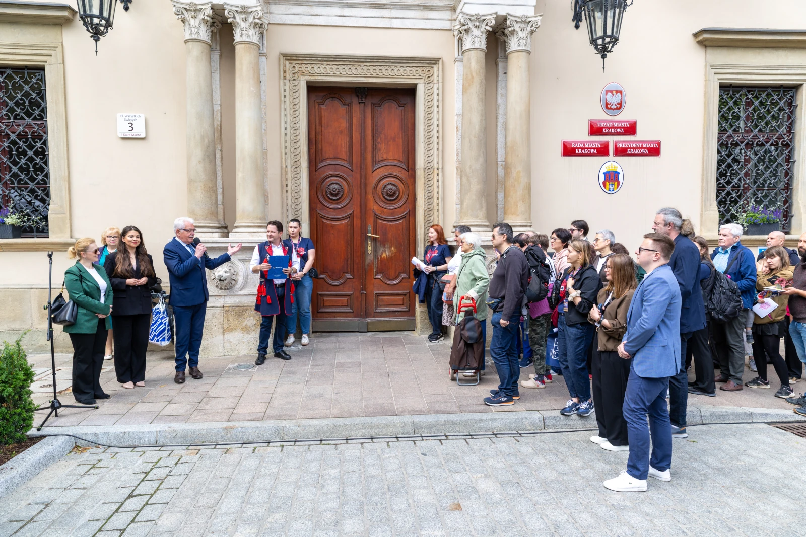 Grupa ludzi słuchająca przemówienia przed budynkiem z ozdobnymi drzwiami.  Autor: Bogusław Świerzowski