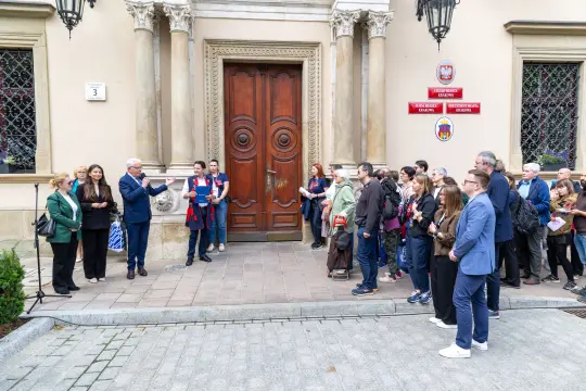 Grupa ludzi słuchająca przemówienia przed budynkiem z ozdobnymi drzwiami.