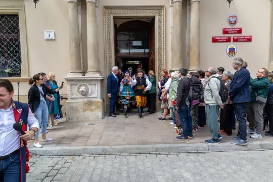 Grupa ludzi w tradycyjnych strojach, witająca gości przed budynkiem.