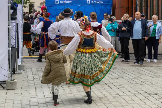 Tradycyjne stroje ludowe w Krakowie, tańczące postacie i tłum widzów.