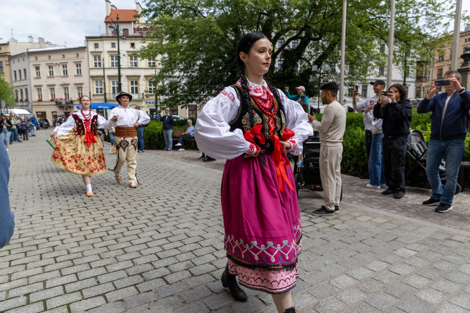 Kobieta w tradycyjnym stroju ludowym idzie ulicą, w tle tańczący ludzie i obserwatorzy.  Autor: Bogusław Świerzowski