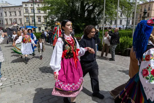 Kobieta w tradycyjnym stroju ludowym idzie w procesji, towarzyszą jej inne osoby w strojach regionalnych.