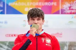 Zawodnik w czerwonym stroju trzyma złoty medal i uśmiecha się.