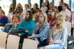 Grupa ludzi siedzących na wykładzie lub prezentacji, różne reakcje i zainteresowanie.