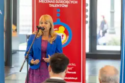 Kobieta przemawia do mikrofonu, w tle plakat promujący mecenat dziecięcych talentów.