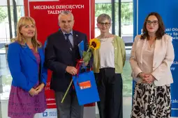 Cztery osoby stojące obok siebie, trzymające kwiaty i torby, w tle czerwone i niebieskie banery.