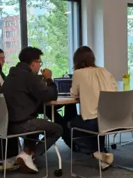 Dwie osoby siedzą przy stole, pracując na laptopie w jasnym pomieszczeniu.
