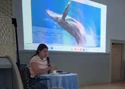Kobieta siedząca przy stole, prezentująca zdjęcie skaczącego wieloryba na ekranie.