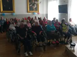 Grupa osób starszych na wózkach inwalidzkich w sali, uczestniczących w wydarzeniu.