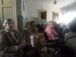 Grupa osób starszych w sali, niektórzy na wózkach inwalidzkich, uczestniczy w wydarzeniu.