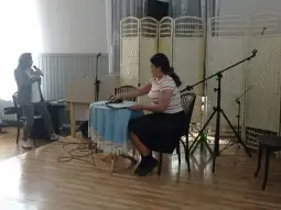 Kobieta przy stole z laptopem, druga osoba śpiewa na scenie.