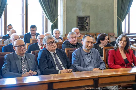 Grupa osób siedzących w sali obrad, wśród nich mężczyźni w garniturach i kobieta w czerwonej marynarce.