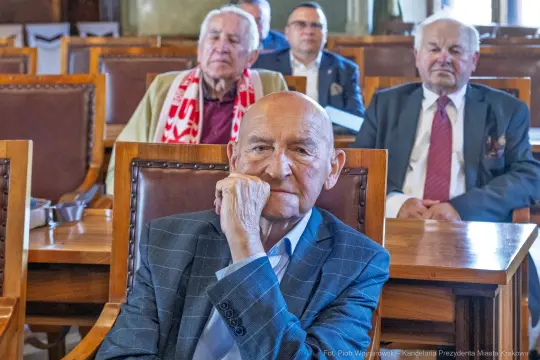 Mężczyzna w garniturze siedzi w sali, w tle inni uczestnicy spotkania.