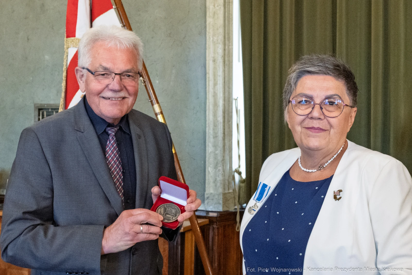 Mężczyzna wręcza medal kobiecie w eleganckich strojach, w tle flagi i zasłony.  Autor: Piotr Wojnarowski
