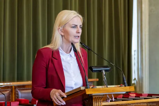 Kobieta w czerwonym żakiecie przemawia z mównicy w sali obrad.