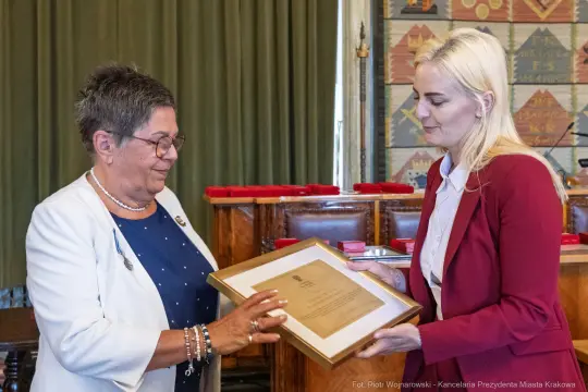 Wręczenie nagrody przez kobietę w białej bluzce drugiej kobiecie w czerwonej marynarce.