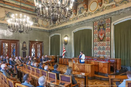 Sala obrad z uczestnikami, mówiąca kobieta przy mównicy, dekoracyjne detale architektoniczne.